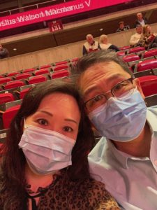 An Asian American woman and man wearing disposable surgical face masks while sitting inside a large stadium.